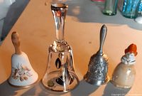 Photo of four decorative bells showing different materials and designs: crystal, porcelain with painted flower and wooden handle, embossed silver-tone metal, and ceramic hen-shaped bell.