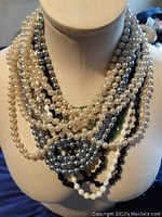 Multiple strands of pearl style necklaces in white, silver, black, and green displayed on a mannequin showing layering and clasps.