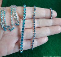 Photo of a hand holding two tennis bracelets and earrings, showing the size and details of light blue rhinestones set in silver-tone metal.