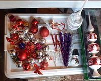 Photo showing a tray containing various Christmas balls mainly in red and silver colors, small gift box ornaments, red bows, purple twisted icicles, and a clear icicle decoration.