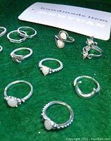 Multiple handmade silver toned rings with white stones and various designs on green surface.