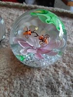 Clear glass spherical paperweight featuring two orange and black honeybee figurines with translucent wings sitting above a pink flower inside a textured glass base with small bubbles, accented by a green swirl leaf inside the top of the sphere.