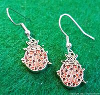 Photo shows pair of silver-tone metal ladybug earrings with detailed First Nations inspired carved patterns, painted red and black, on green fabric background.