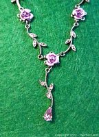 Close-up image of the necklace showing light pink rose decorations, silver leaf designs, and sparkling teardrop stone pendant hanging at the bottom.