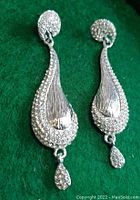 Pair of long silver-tone earrings featuring textured metal and rhinestones on a green background.