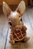 Front view of a small rabbit figurine holding a detailed red and yellow picnic basket with blue eyes and large ears.