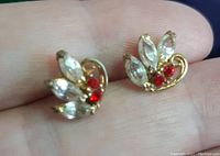 Close-up showing one earring with clear marquise-shaped and round red stones on a gold plated base held between thumb and forefinger.
