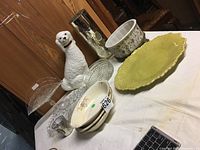 Wide view showing grouped lot on table with ceramic dog figurine, yellow plate, clear glass dishes, and floral pattern dishes.