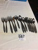 Flatware laid out on white cloth showing 2 serving forks, 2 dinner forks, 2 dessert forks, 8 spoons, 1 sugar spoon, and 1 butter knife with floral patterned handles.