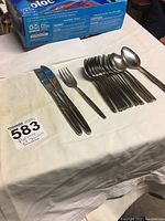 Photo showing two cutting knives, one fork, two serving spoons, and a group of teaspoons arranged on a white cloth.