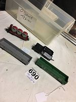 Four HO gauge train cars on a white cloth with a container labeled 'O gauge trains' in the background. The cars include a gray boxcar, black coal tender, green cargo car, and a flatcar with three cylindrical loads.