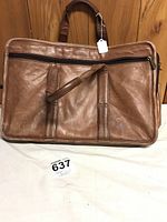 Front view of brown leather briefcase standing upright showing dual top handles and front zippered pocket.
