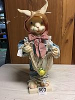 Motorized bunny figurine standing on a white square base, wearing bonnet, cloth clothing, holding fabric pouch with decorative eggs