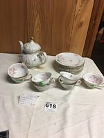 Photo showing teapot, seven cups, and eight plates with white porcelain, pink floral design, and gold accents on a cloth background.