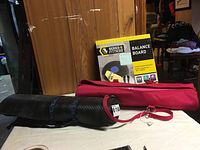 Photo showing pink Gaian yoga mat in carry case, black thick yoga mat with straps, and Series 8 Balance Board in box on table.