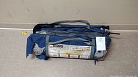 Blue and gray backpacking tent packed inside matching blue carry bag with gray straps, showing seller lot tag.