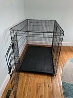 Photo showing the entire black metal dog crate on a hardwood floor with removable plastic tray visible at the bottom and one door open.