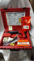 Closed red plastic carrying case open showing all kit contents including the powder-actuated tool, Ramset fasteners box, cartridge pack, cleaning brushes, and paper manuals inside fitted compartments.