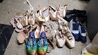 Overview of all dance shoes showing multiple pairs of nude satin pointe shoes, a pair of black leather shoes, and a pair of colorful rainbow pattern shoes.