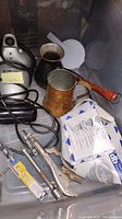 Photo showing copper cup, copper pot with wooden handle, Snap-on locking pliers, metal keychain tool, metal rod with attachment, small electric appliance with power cord, and metal sink basket in plastic bin.