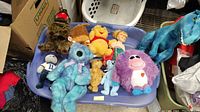 Top view of 11 assorted stuffed animals in blue container including Winnie the Pooh, dinosaur, and purple furry plush