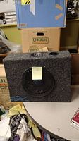 Full view of The Box speaker on a table surrounded by moving boxes and cloths, showing the carpeted enclosure and speaker grille.