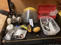 Safety gear and hardware items on a black table: face shield, ear protectors, respirator, masks, safety glasses, and totes with cabinet hardware.