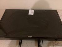 Samsung flat screen TV on a flat surface with a lot number sticker on the screen. The TV has a black bezel and Samsung branding on the front center.