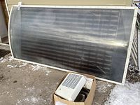First image shows large rectangular curved solar panel with a white frame sitting on a concrete surface outdoors, with a cardboard box containing a fan unit in front.