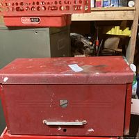 Front view of red metal tool chest showing closed top lid, white handle, and a badge on the front.