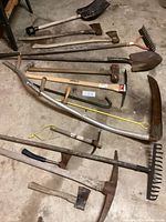 Photo showing full assortment of 15 used garden and hand tools including multiple rakes, shovels, axes, a pick, a scythe, and other tools laid out on a concrete floor.