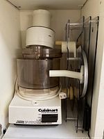 White Cuisinart DLC-10E food processor body with translucent mixing bowl and attached accessories stored upright behind it.