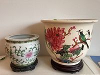 View of two ceramic planters of different sizes on their wood bases, showing floral and bird designs