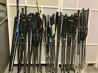 Photo showing a large number of golf shafts leaning against a white background. Shafts have various grips including black grips with Ping branding and Golf Pride grips. Mizuno Shaft Optimizer practice drivers with blue grips are visible.