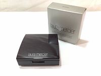 Closed compact powder case with outer silver box in background, showing laura mercier logo on case and box.