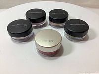 Five jars of makeup products: four BareMinerals loose mineral eye shadows with black lids and one Artdeco lip glitter with silver lid.