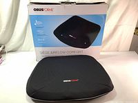 Top front view of the black ergonomic seat cushion placed in front of its box with brand and product information visible.