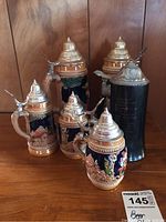 Five German beer steins arranged on a wood surface with detailed ceramic painting and pewter lids.