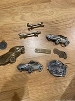 Top-down photo showing all belt buckles on wooden floor, various vehicle and animal shapes and a brass engraved plate.