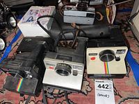 Three black Polaroid instant cameras with rainbow stripes on bottoms, Kodak Instamatic 704 camera with leather strap, and a GE Flashbar II flash unit.