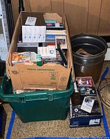 Photo showing a green plastic bin and cardboard box containing cassette tapes, DVDs, and records along with some loose cassettes and eight track tapes around it.