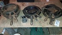 Multiple stacks of cast iron skillets on a wooden table with a small metal pitcher
