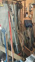 Photo showing assorted chains, garden tools with wooden and metal handles including shovels and rakes organized against a wall in garage space.