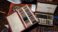 Photo showing two vintage slide storage boxes filled with mounted photographic slides, one box open with an index sheet listing slide details in handwritten text.