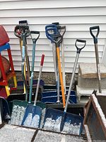 Photo showing a collection of snow shovels and pushers with a variety of handle styles (plastic D-grips, wooden, metal) and blades (plastic and metal edges), some with traces of snow.