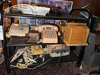 Wide view of a metal cart shelf with multiple vintage telephones including rotary dial, push button, wooden, and beige CB Radio Telephone models.