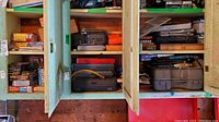 Green wooden cabinet with multiple shelves containing plastic tool cases, boxes, and packaged tools. Visible boxes for air conditioner retrofit kits, staplers, drill bits, and other hardware items.