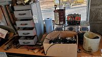 Photo showing metal chest of drawers, cardboard box of various parts, hand tools scattered, and battery charger on surface