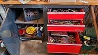 Wide view of red Craftsman tool chest with three drawers open showing assorted tools and small hardware boxes. Black metal shelving unit visible to the left with automotive funnels and battery tester on the shelf.