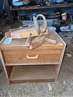 Wooden table with drawer and woodworking vise on top, sitting on a dirt floor in a garage-like space.
